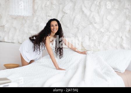 Smiling pretty millennial european brunette woman with long curly hair makes bed with blanket in white bedroom Stock Photo