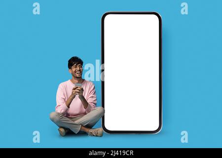 Happy indian guy hipster sitting by big smartphone, mockup Stock Photo