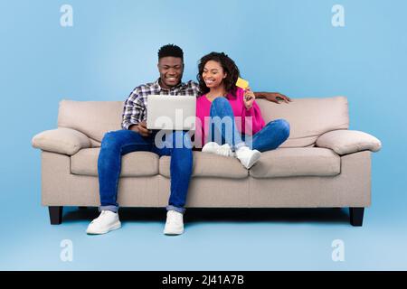 Black couple shopping online via laptop and credit card, ordering things in internet, sitting on sofa, blue background Stock Photo