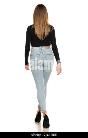 Rear view of a young woman walking in studio in black short blouse and blue jeans on a white background Stock Photo