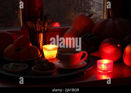 Different types of pumpkins lie on the windowsill, candlesticks with burning candles, as well as a cup of black tea, pumpkin seed cookies on a saucer Stock Photo