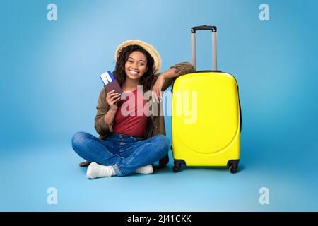 Lovely young black woman holding passport and plane tickets, sitting near bright yellow suitcase on blue background Stock Photo