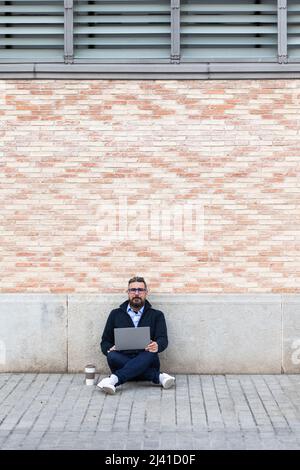 Man sitting on sidewalk and leaning against wall using his laptop while looking at camera Stock Photo