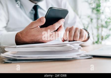 Businessman searching information online while using smartphone over paperwork and report stack, selective focus Stock Photo