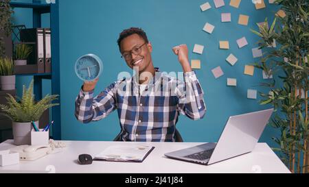 Happy employee checking time on clock and finishing work before deadline, doing countdown for timeout. Punctual man feeling cheerful about break timing, looking at watch with hour and minutes. Stock Photo