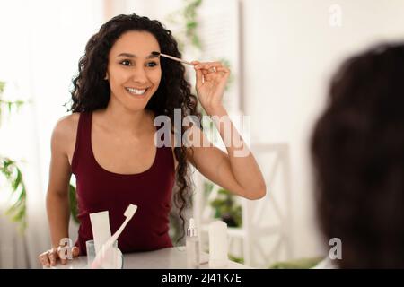 Charming millennial woman applying eyebrow mascara near mirror at home, empty space Stock Photo