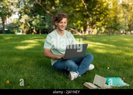 Cheerful caucasian male student using laptop and learning online, doing homework outdoors, sitting on lawn Stock Photo
