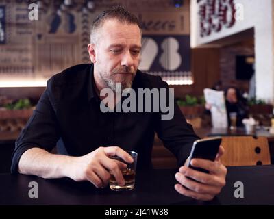 Caucasian middle age man holding a glass of whiskey sits at the bar counter and looks at his mobile phone. Stock Photo