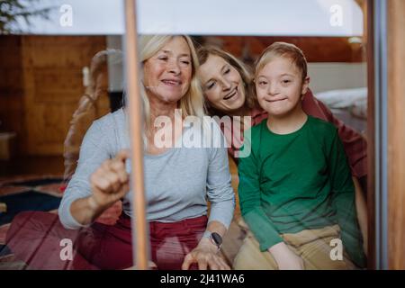Boy with Down syndrome with his mother and grandmother sitting and looking through window at home. Stock Photo