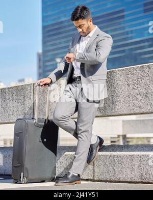 I hope Im not going to be late. Shot of a young businessman checking the time while standing with a suitcase in the city. Stock Photo