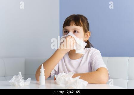 Little girl with runny nose is blowing into paper tissue. Nasal spray and used tissues on table Stock Photo