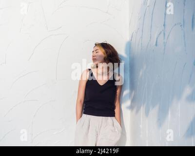 Attractive Asian woman portrait with short hair wearing black tank top and white trousers standing and keep hands in pockets on white wall background, Stock Photo
