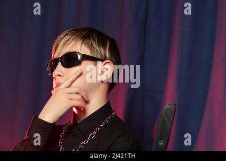 Portrait of a stylish teenager 13 years old with a fashionable hairstyle in black sunglasses with a chain around his neck on a purple background. Sele Stock Photo