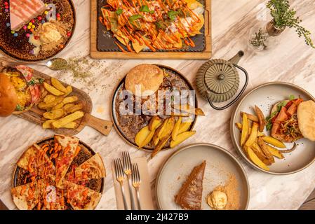 Set of fast homemade food dishes with assorted burgers, nachos with guacamole and nacho cheese, homemade fries, assorted cakes on marble table Stock Photo