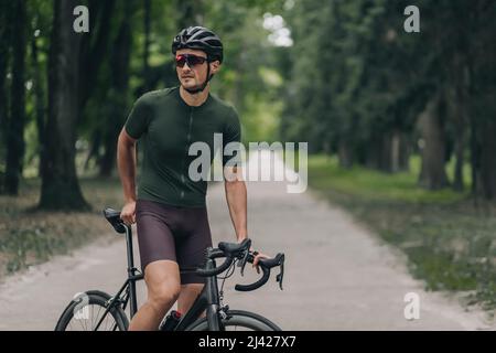 Strong young man sitting on bike during recreation after outdoors training. Professional cyclist wearing sport clothes, protective helmet and mirrored Stock Photo