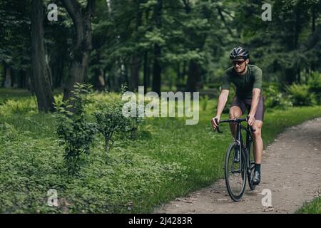 Muscular young man wearing sport clothes, protective helmet and mirrored glasses riding bike at green city park. Outdoors activity of caucasian guy. H Stock Photo