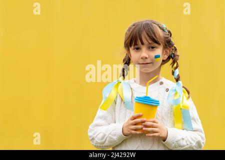 A little Ukrainian girl with two pigtails and yellow and blue ribbons drinks a hot drink from a glass of patriotic colors like the Ukrainian flag. Stop the war in Ukraine. Volunteer Help for Refugees. High quality photo Stock Photo