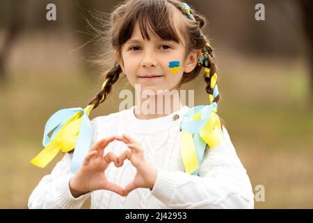 A beautiful little girl with pigtails and braided yellow and blue ribbons, like the color of the Ukrainian flag, makes a heart with her hands. I love Ukraine. Stop the war. . High quality photo Stock Photo