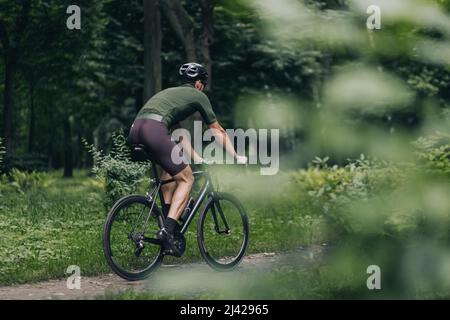 Back view of sporty young man in activewear and helmet cycling outdoors. Male athlete spending leisure time for sport activity at summer park. Stock Photo