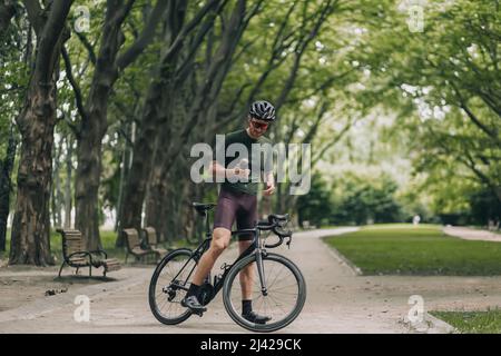 Full length portrait of tired cyclist sitting on black bike and holding sport bottle of fresh water. Caucasian man wearing safety helmet and mirrored Stock Photo