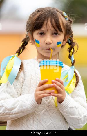 A little Ukrainian girl with two pigtails and yellow and blue ribbons drinks a hot drink from a glass of patriotic colors like the Ukrainian flag. Stop the war in Ukraine. Volunteer Help for Refugees. High quality photo Stock Photo