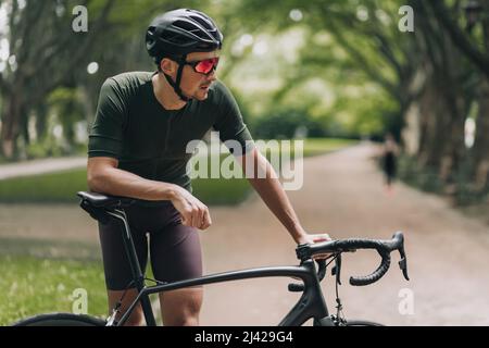 Side view of sporty young man leaning on bike while taking break during morning ride at city park. Caucasian cyclist wearing sport clothes, safety hel Stock Photo