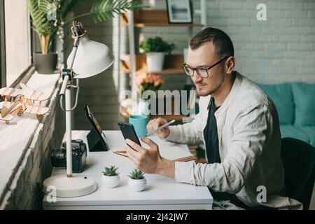 Entrepreneur businessman working sitting at desk holding smartphone writing in diary with pen, using tablet pc. Businessman manager employee Stock Photo