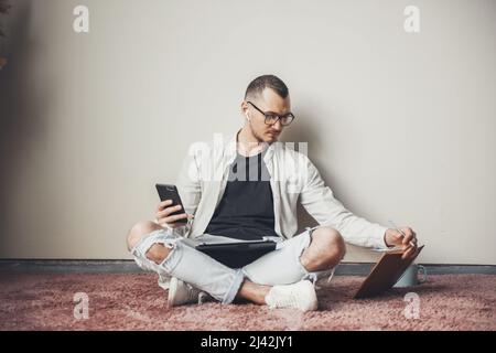 Student man using mobile smart phone, looking at his textbook browsing internet application on smartphone holding digital tablet, sitting on the floor Stock Photo