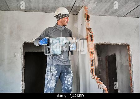 Side view of man builder in workwear drilling wall with hammer drill. Male worker using drill breaker while destroying wall in apartment under renovation. Demolition work and home renovation concept. Stock Photo