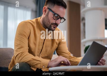 Shot of focused adult man, working online on laptop, sitting in his living room. Stock Photo
