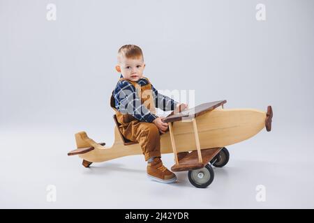 A 3-year-old boy in a checkered shirt sits on a large wooden plane. Children's environmentally friendly toys from wood Stock Photo
