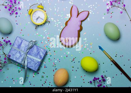 Festive Easter background top view. Easter bunny, pastel colored painted eggs, purple flowers, paint brush, gift, box, retro yellow alarm clock lie am Stock Photo