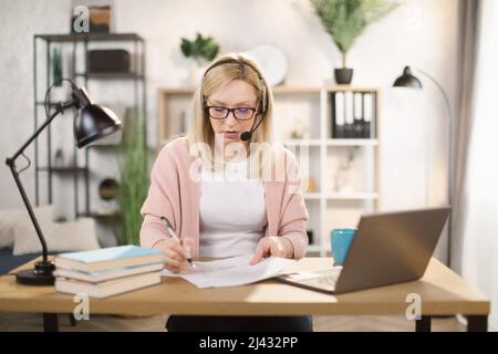 Smiling Caucasian woman writing notes while using laptop computer at bright office or home interior. Pretty female software developer using modern laptop for remote work at home. Stock Photo