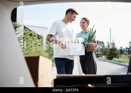 Portrait of smiling adult couple loading boxes in car trunk while moving into new house scene in sunlight Stock Photo