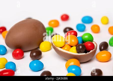 A whole and half of a cracked chocolate egg filled with multi-colored candies on a white background with multi-colored candies, copy space. Treats for Stock Photo