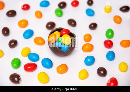 Half of a cracked chocolate egg filled with multi-colored candies on a white background with multi-colored candies, top view. Treats for children. Swe Stock Photo