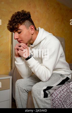 Caucasian young man with curly hair sitting on his bed with a worried gesture, he analyzes the problems that the step of becoming an adult man entails Stock Photo