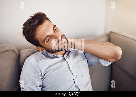 Hes feeling a bit of tension on his neck. Shot of a handsome young man holding his neck in pain while relaxing on a sofa at home. Stock Photo