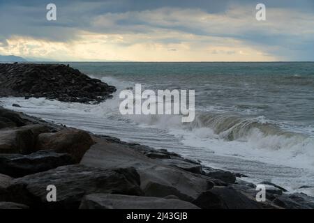 Storm waves roll over large rocks on the seashore with dark clouds and a clear sun in the sky Stock Photo