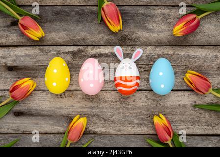 Colorful Easter eggs, one egg with cute bunny face and rabbit ears next to orange-yellow tulips on a rustic wooden table. Easter concept. Stock Photo