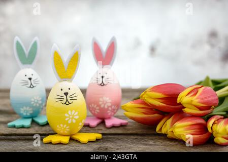 Three Easter eggs with cute bunny face and rabbit ears next to orange-yellow tulips on a rustic wooden table. Easter concept. Stock Photo