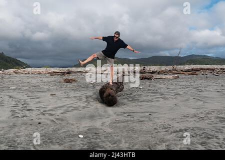 Testing his balance, an older man stands on one leg on a driftwood log on the beach, he's having fun Stock Photo