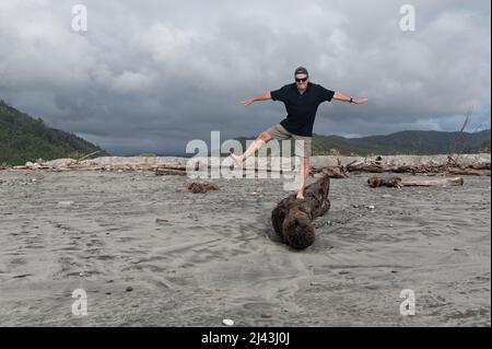 An older man is balancing on one leg while standing on a log on the beach Stock Photo