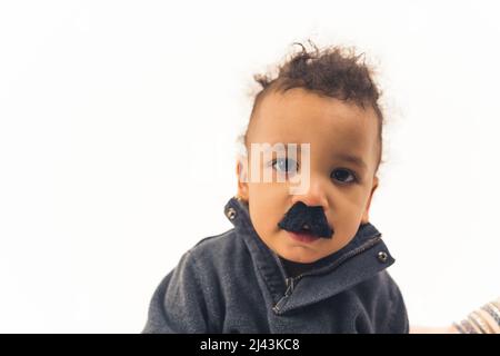 little African American curly boy with big black eyes and toy mustache looking into the camera studio shot medium closeup white background copy space. High quality photo Stock Photo