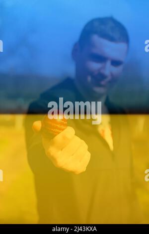 Defocus ukrainian young man holding one colored red egg on nature background. Easter, Ukraine flag. Craft painted eggs. Pysanka or krashanka. Out of f Stock Photo
