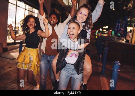 Cheerful young woman piggybacking her friend in the city. Girlfriends cheering and having fun while going out with their friends at night. Group of vi Stock Photo