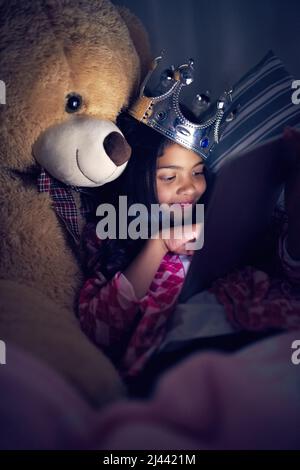 Bedtime stories have never been more exciting. Cropped shot of a little girl using a digital tablet while lying in bed with her teddy at night. Stock Photo