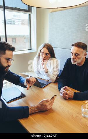 Colleagues working together in a modern co-working space. Three businesspeople using a smartphone during an important discussion. Team of diverse entr Stock Photo