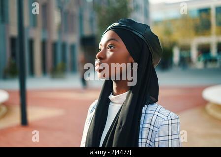 Thoughtful African American female in stylish formal outfit and Muslim scarf observing park with interest and curiosity Stock Photo