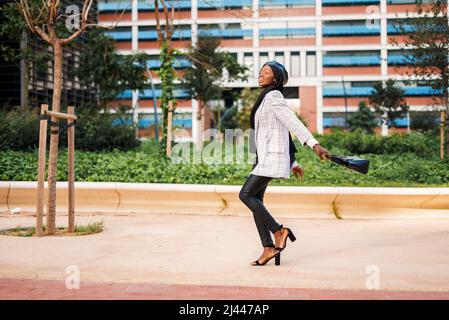 Full body side view of cheerful African American female in stylish outfit with traditional hijab carrying handbag while strolling in park in modern city Stock Photo